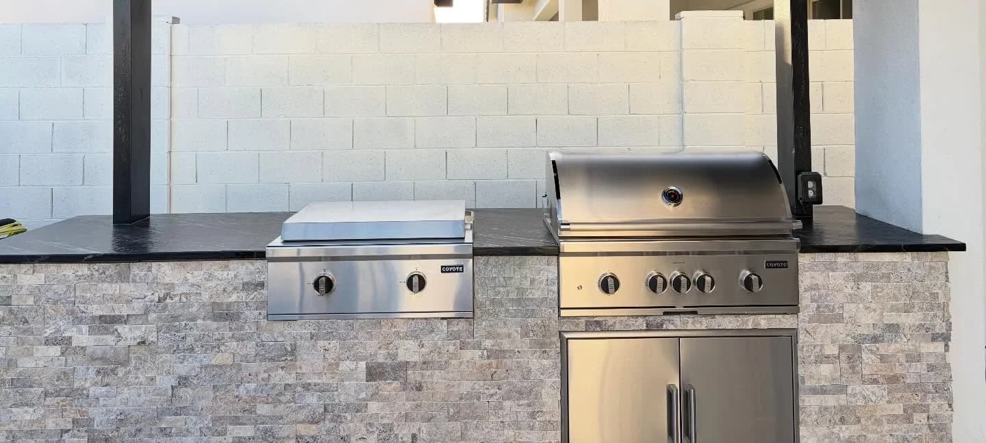 A BBQ island seen straight on: stacked stone veneer, a black stone countertop, a built-in grill with access doors below it and a side burner to its left