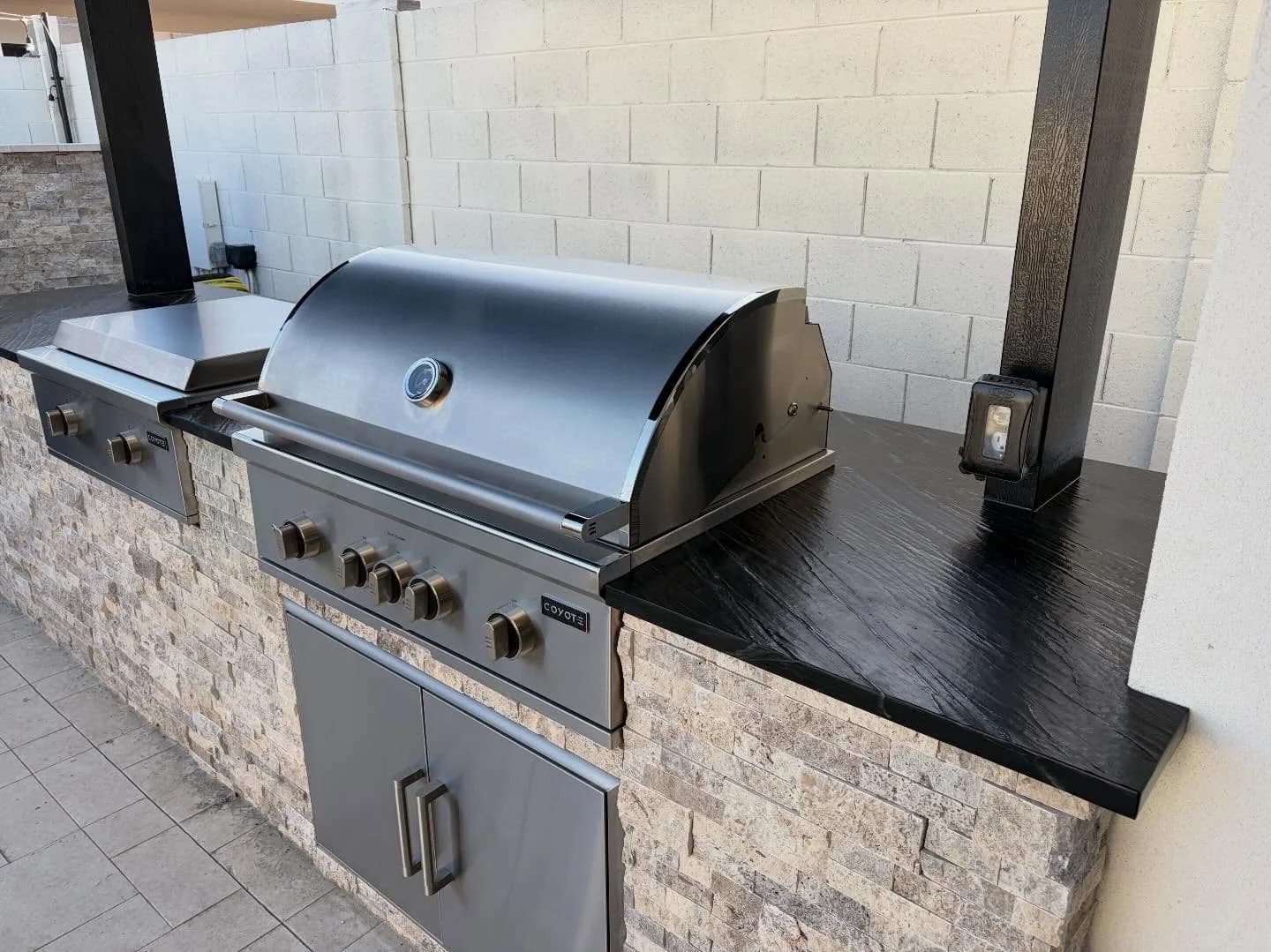 A built BBQ island clad in stacked stone veneer, with a stainless built-in grill, a side burner and access doors under a black stone countertop