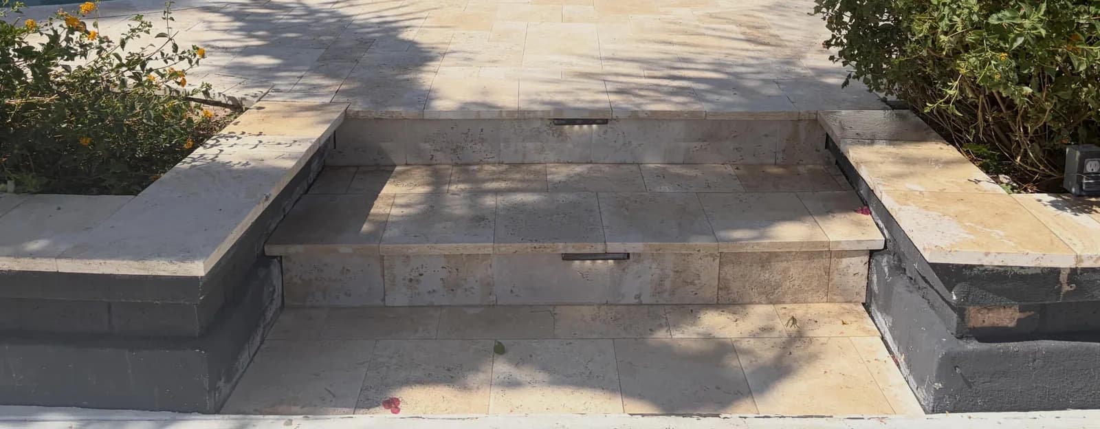 Two raised planters with travertine caps holding orange lantana, one on each side of a short flight of travertine steps